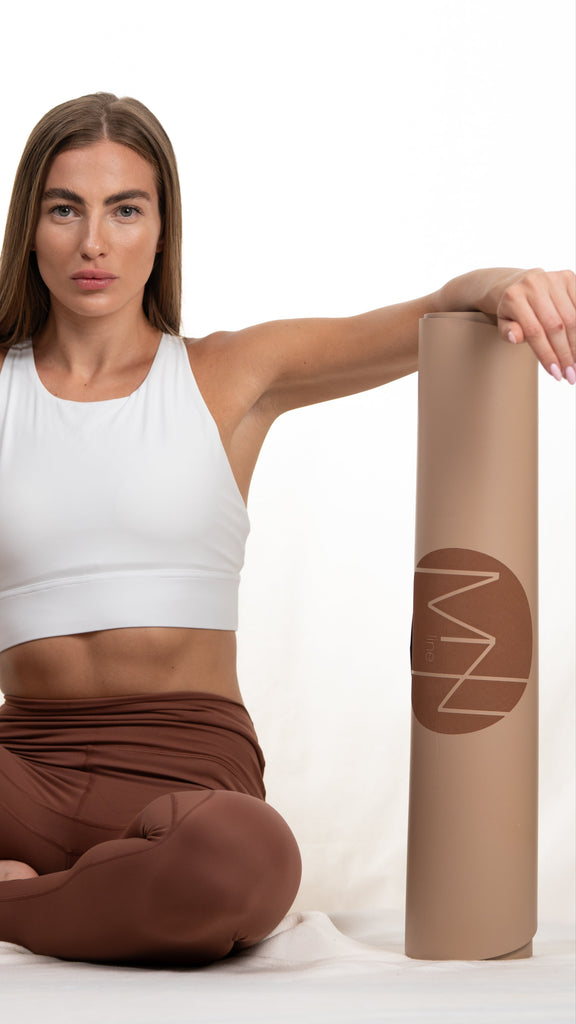 A person sitting on a beige yoga mat with the Mzline logo, wearing a white tank top and brown pants.